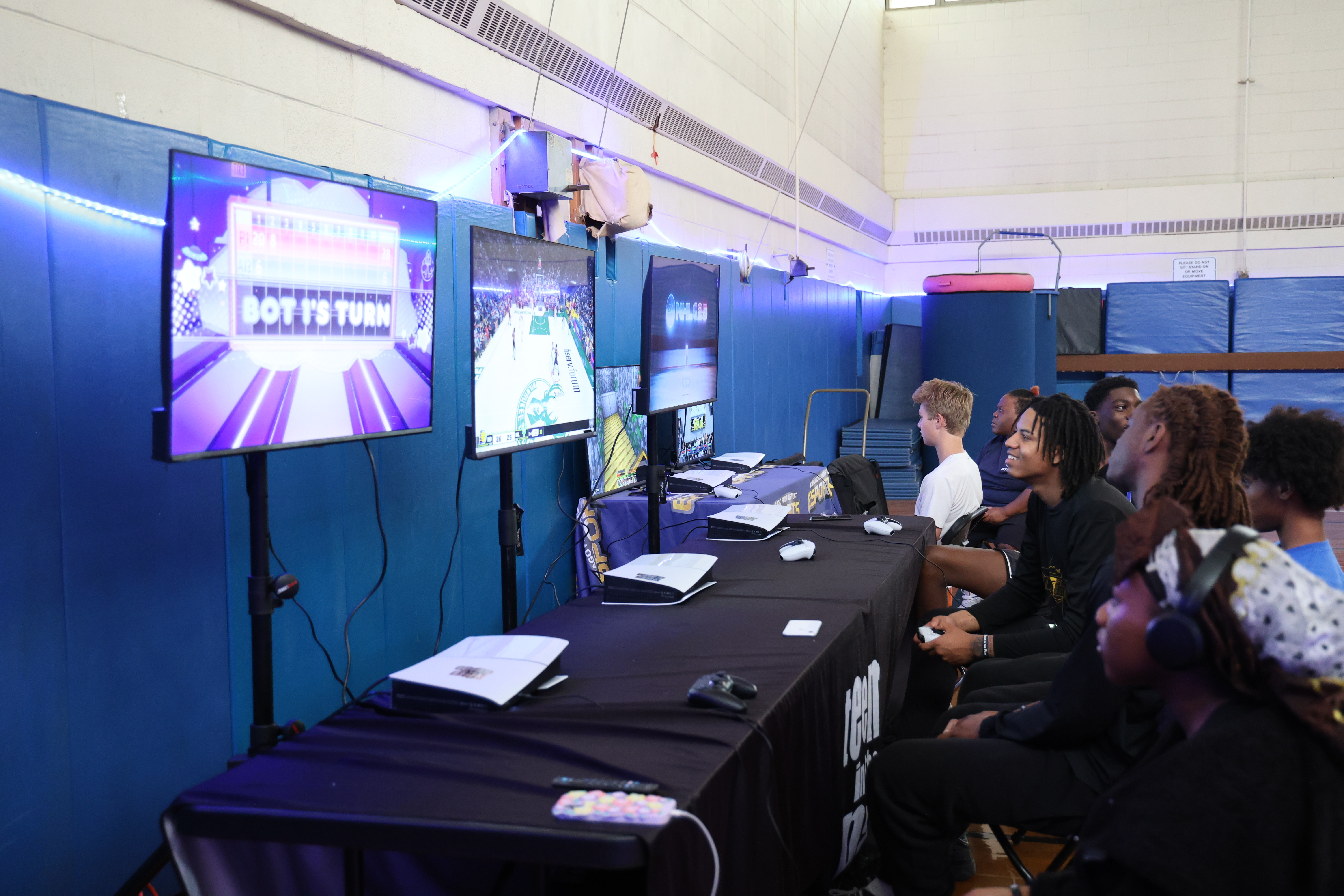 Gamers play video games at a long table with multiple consoles and screens.